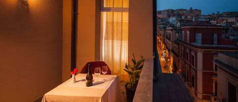 A balcony with a table set for two, a red chair, a potted plant, and a city view at dusk.