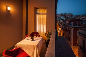 A balcony with a table set for two, a red chair, a potted plant, and a city view at dusk. - Hotel Italia (Cagliari)