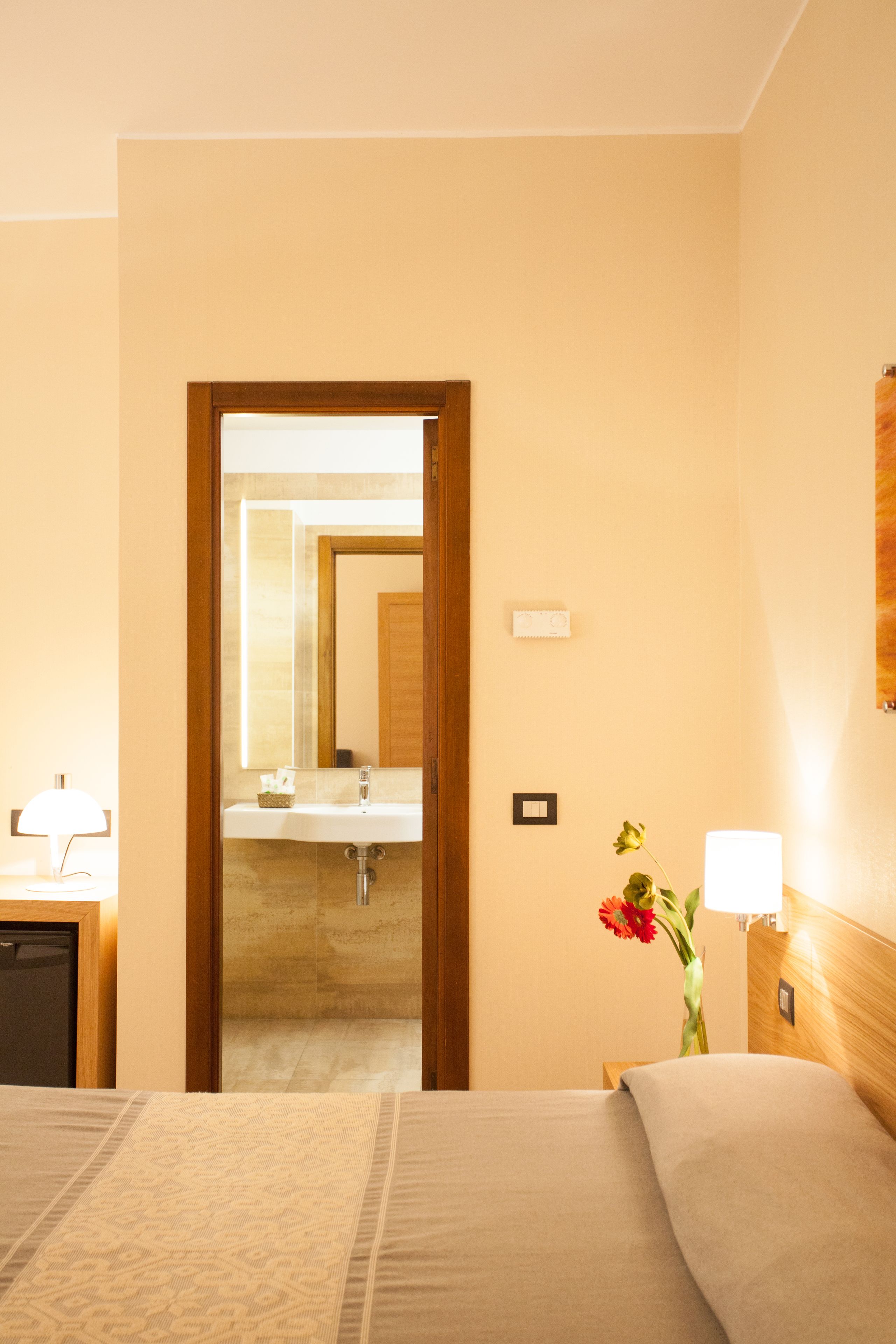 Una camera d'albergo con un letto, un comodino con una lampada, uno specchio, una testiera in legno e un bagno visibile attraverso una porta aperta.