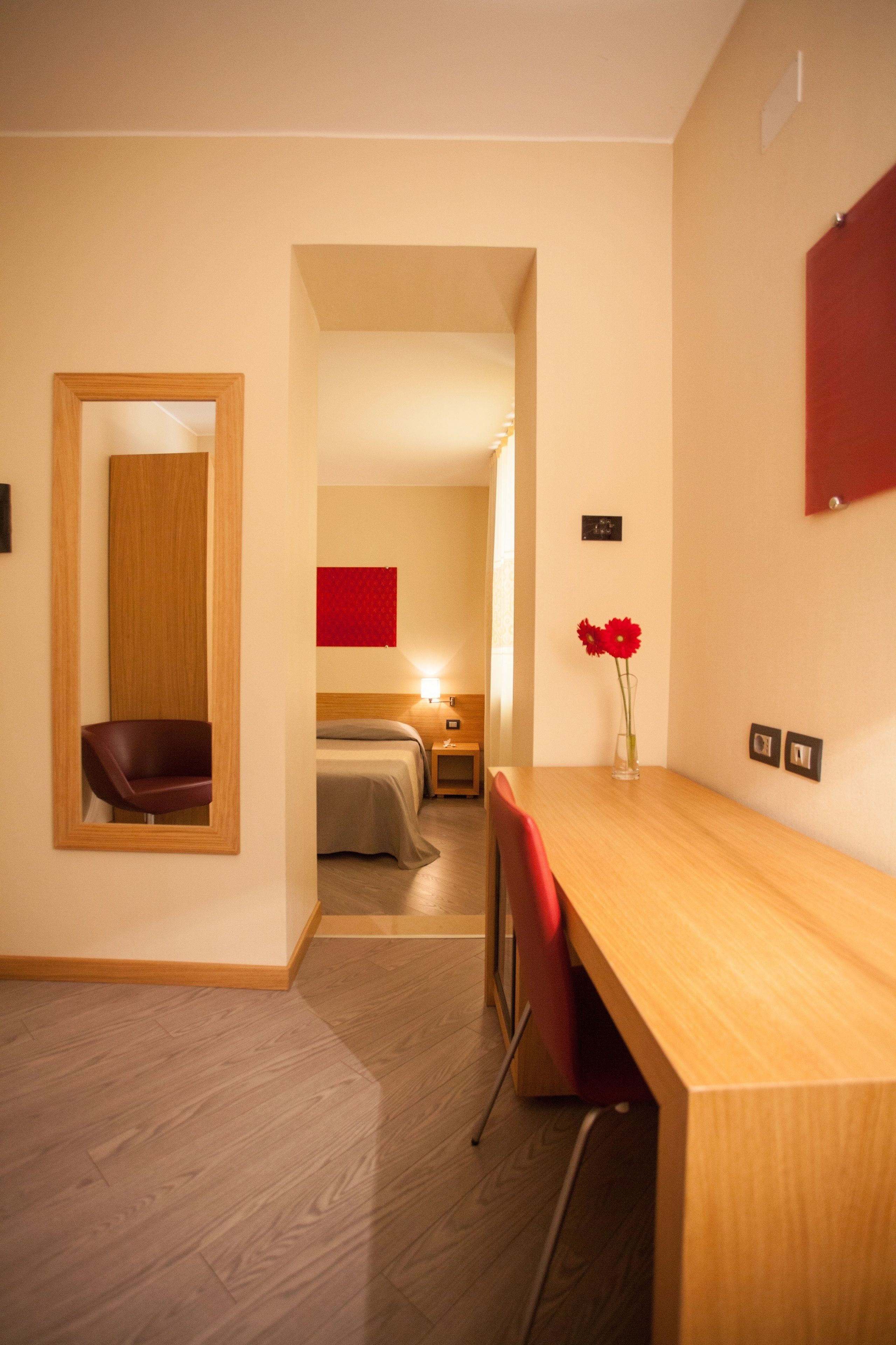 A hotel room with a wooden desk, a red chair, a bed with a wooden headboard, and a mirror on the wall.