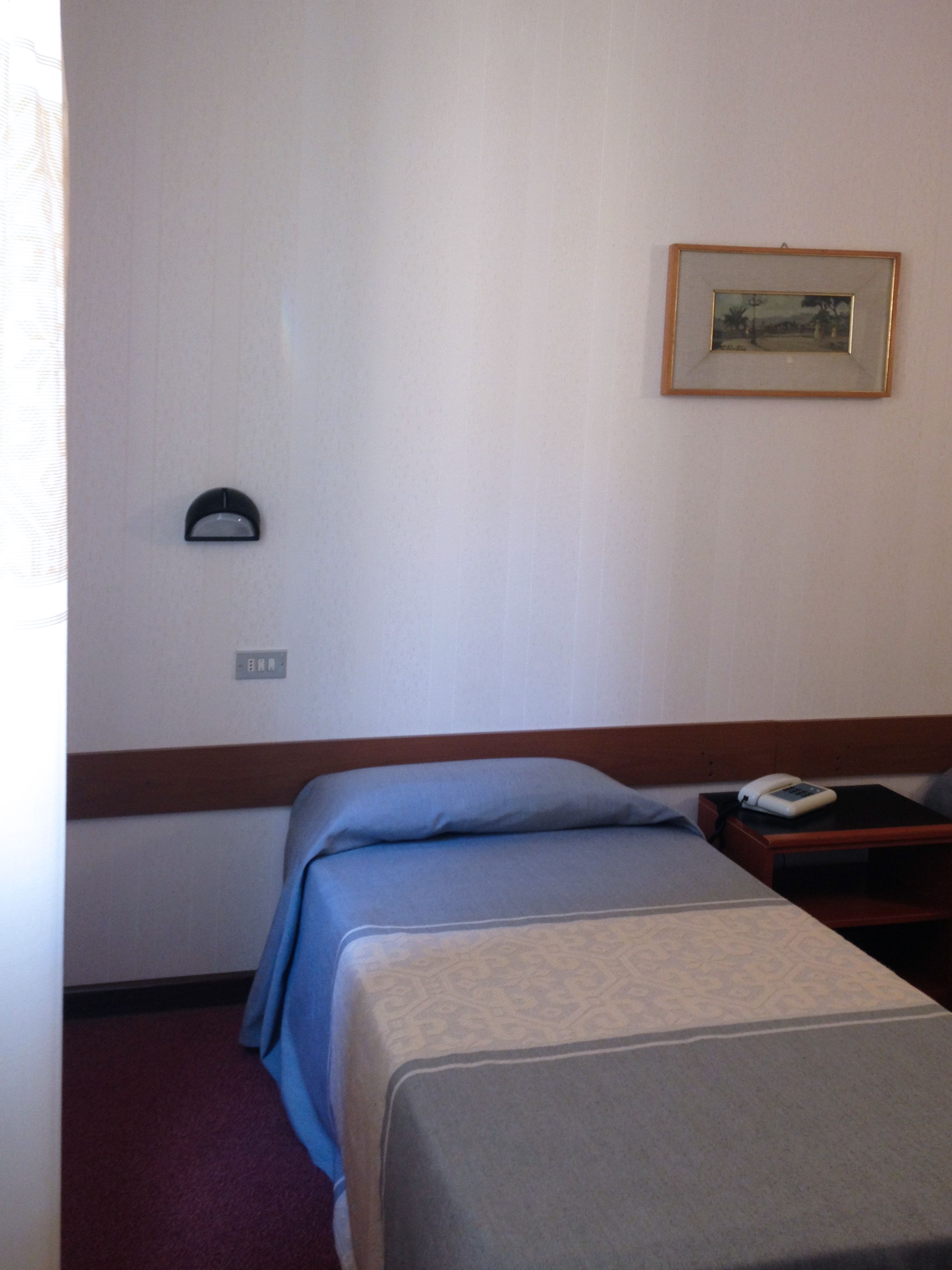 Une chambre d’hôtel avec un lit simple, une table de chevet avec un téléphone, un tableau encadré au mur et une lampe fixée au mur.