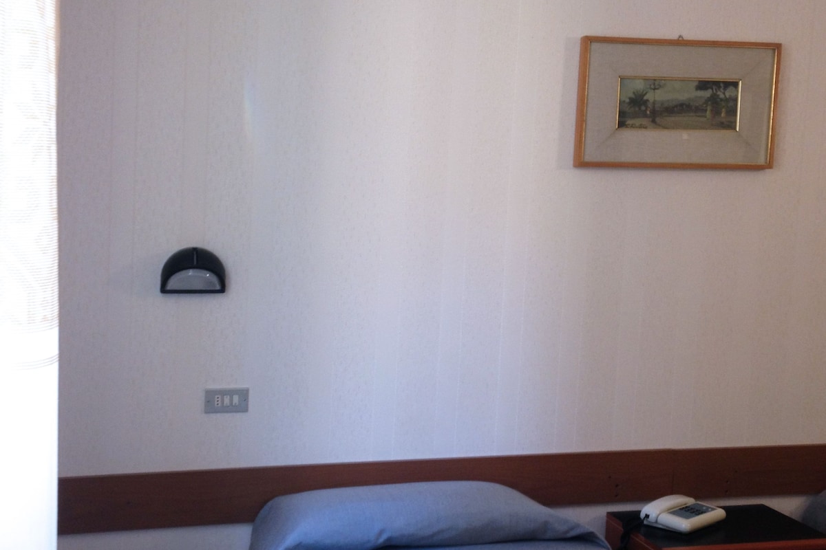 A hotel room with a single bed, a nightstand with a phone, a framed picture on the wall, and a wall-mounted light.