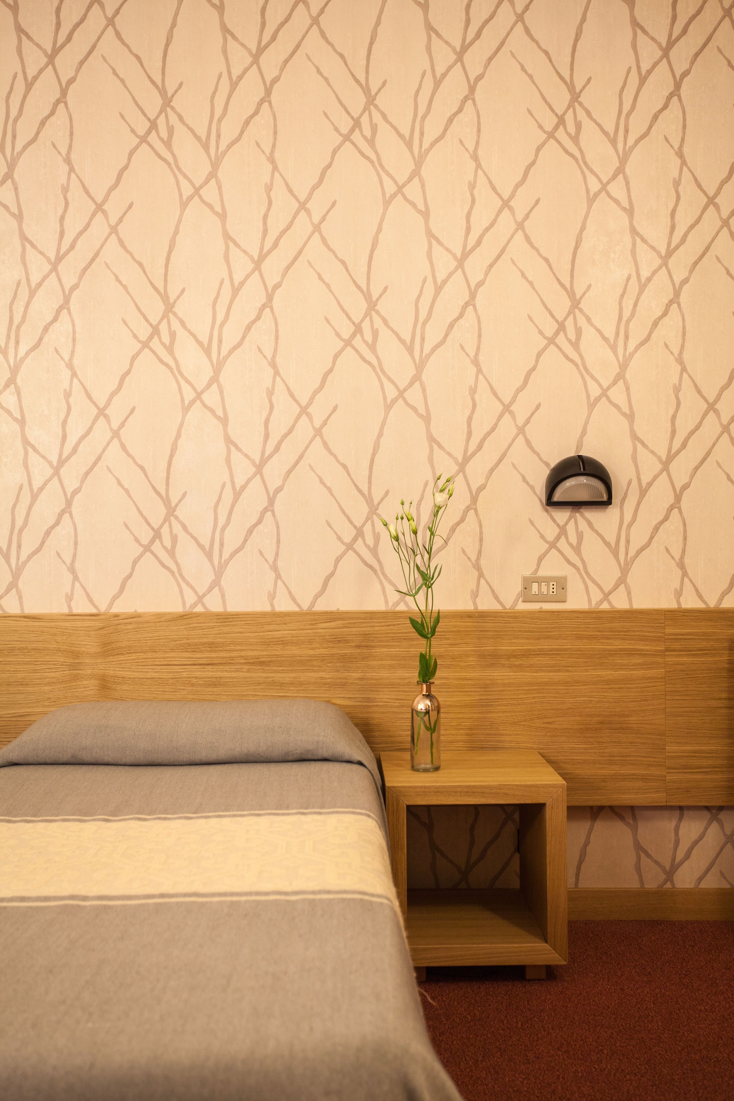A hotel room with a bed, a nightstand with a vase, and a wall-mounted light.