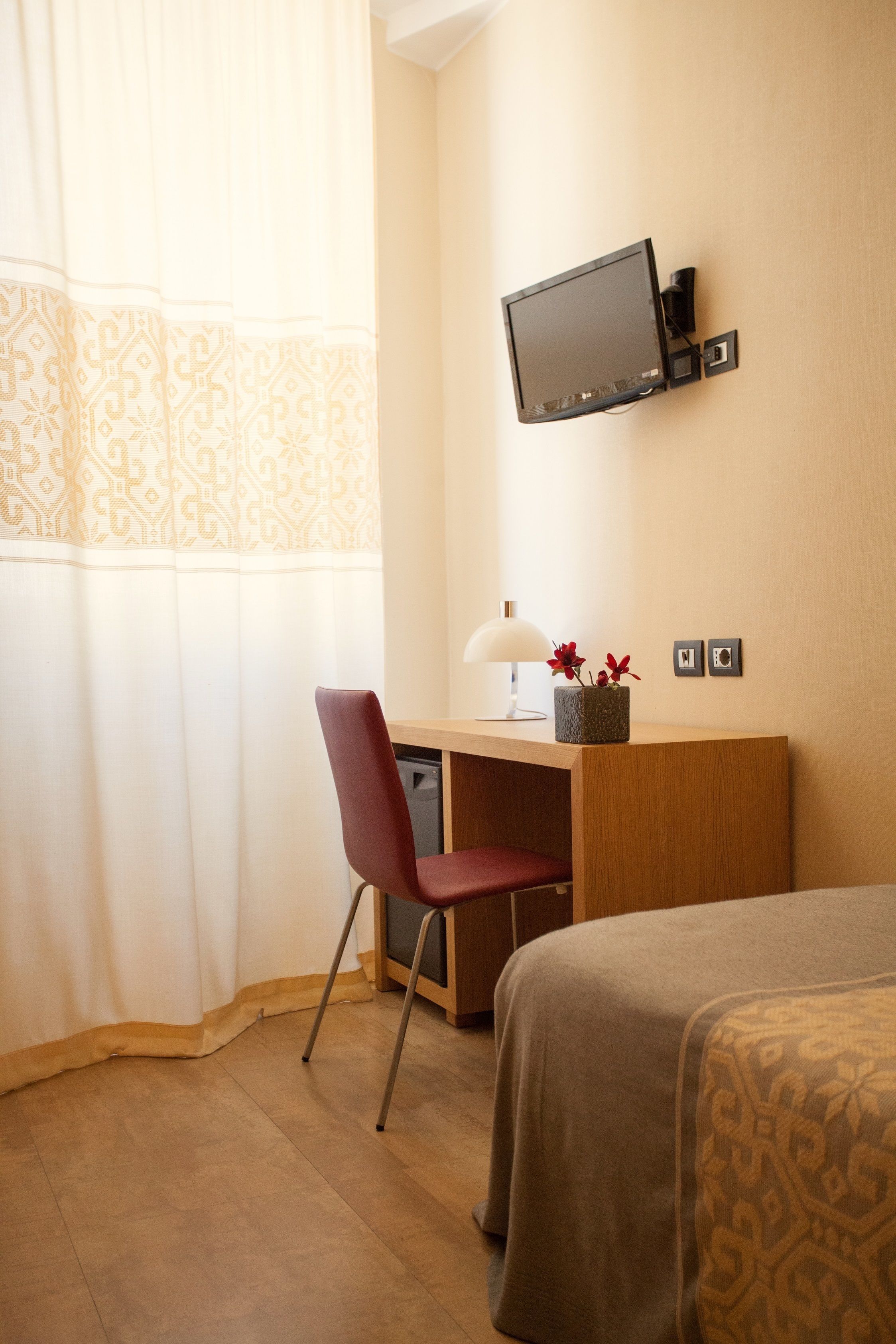 A hotel room with a wooden desk, a red chair, a bed with a patterned bedspread, a mounted TV, and a lamp on the desk.