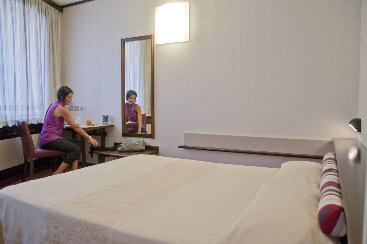 A hotel room with a bed, a desk, a chair, a mirror, and a window with curtains.