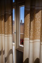 Una ventana con cortinas estampadas, vista de un edificio y un balcón con flores rojas.