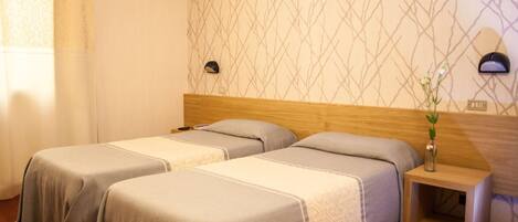 A hotel room with two single beds, a wooden headboard, a nightstand with a vase, and a wall-mounted lamp.