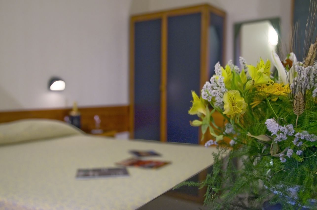 A hotel room with a bed, a bedside table with a lamp, and a floral arrangement on the table.