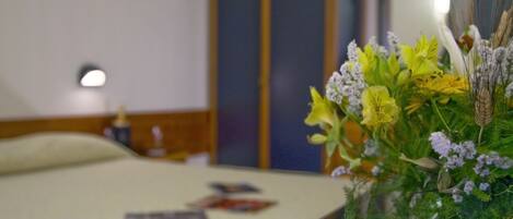 A hotel room with a bed, a bedside table with a lamp, and a floral arrangement on the table.