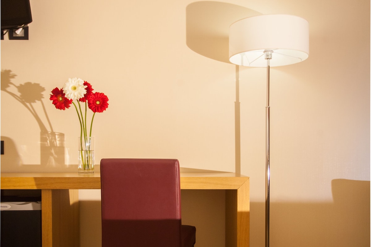 A hotel room with a wooden desk, a red chair, a floor lamp, and a vase with flowers.