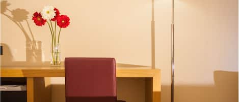 A hotel room with a wooden desk, a red chair, a floor lamp, and a vase with flowers.