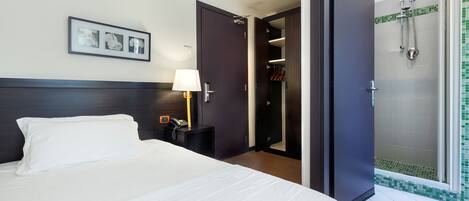 Habitación de hotel con una cama, una lámpara de noche, un teléfono, un cuadro en la pared y una puerta que da a un baño con ducha.