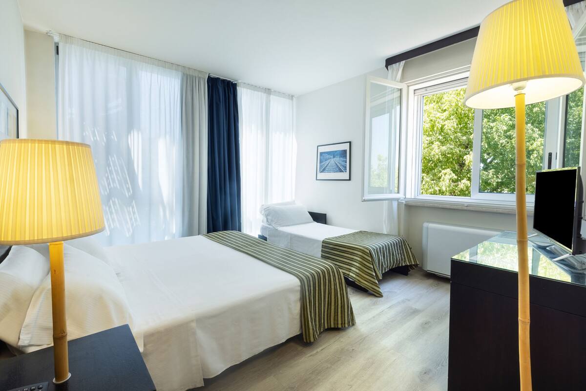 Hotellihuone, jossa on kaksi sänkyä, työpöytä tietokoneella, televisio ja ikkuna, josta on näkymä puita kohti.