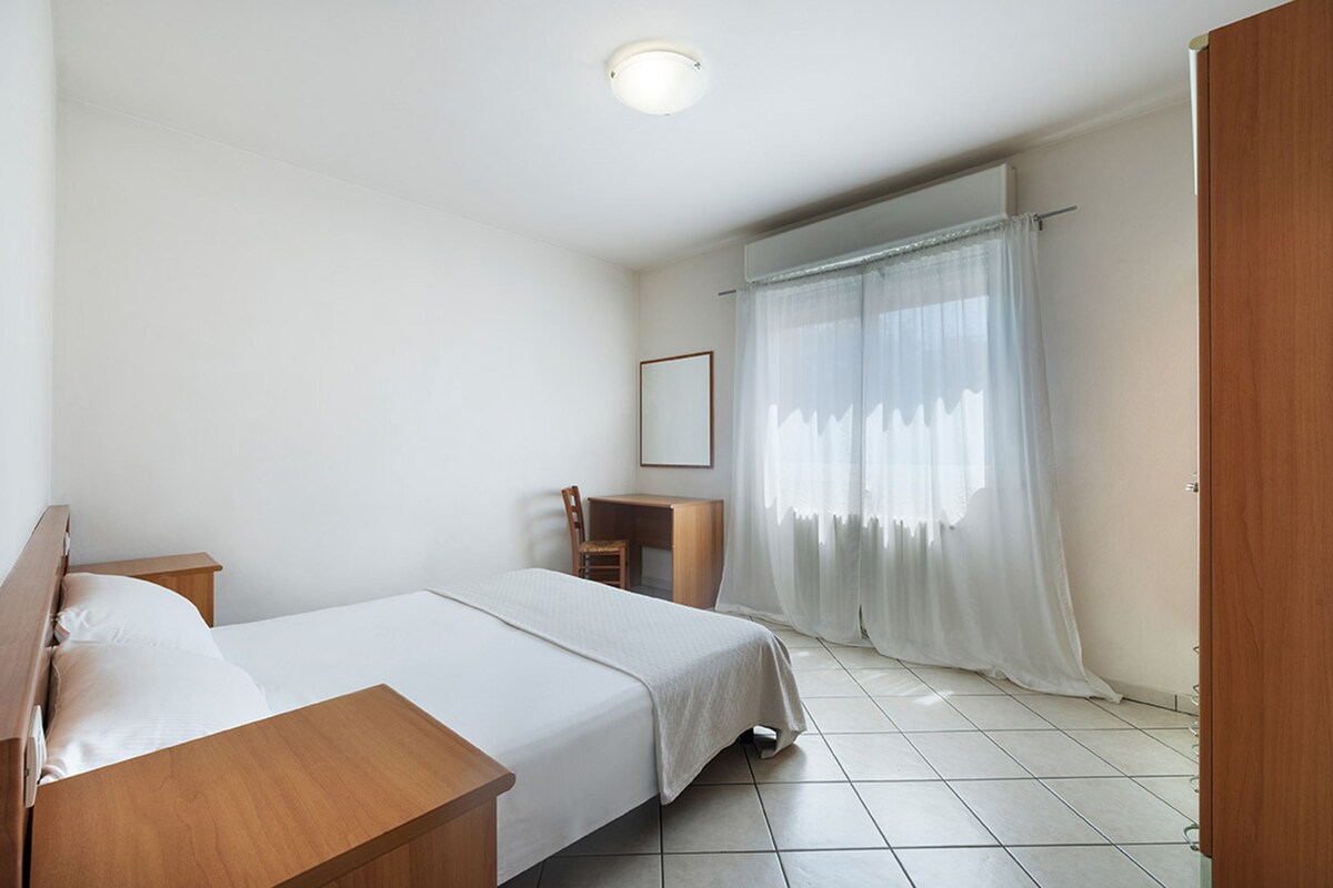 Une chambre avec un lit simple, une table de chevet en bois, un bureau avec une chaise et une fenêtre avec des rideaux légers.