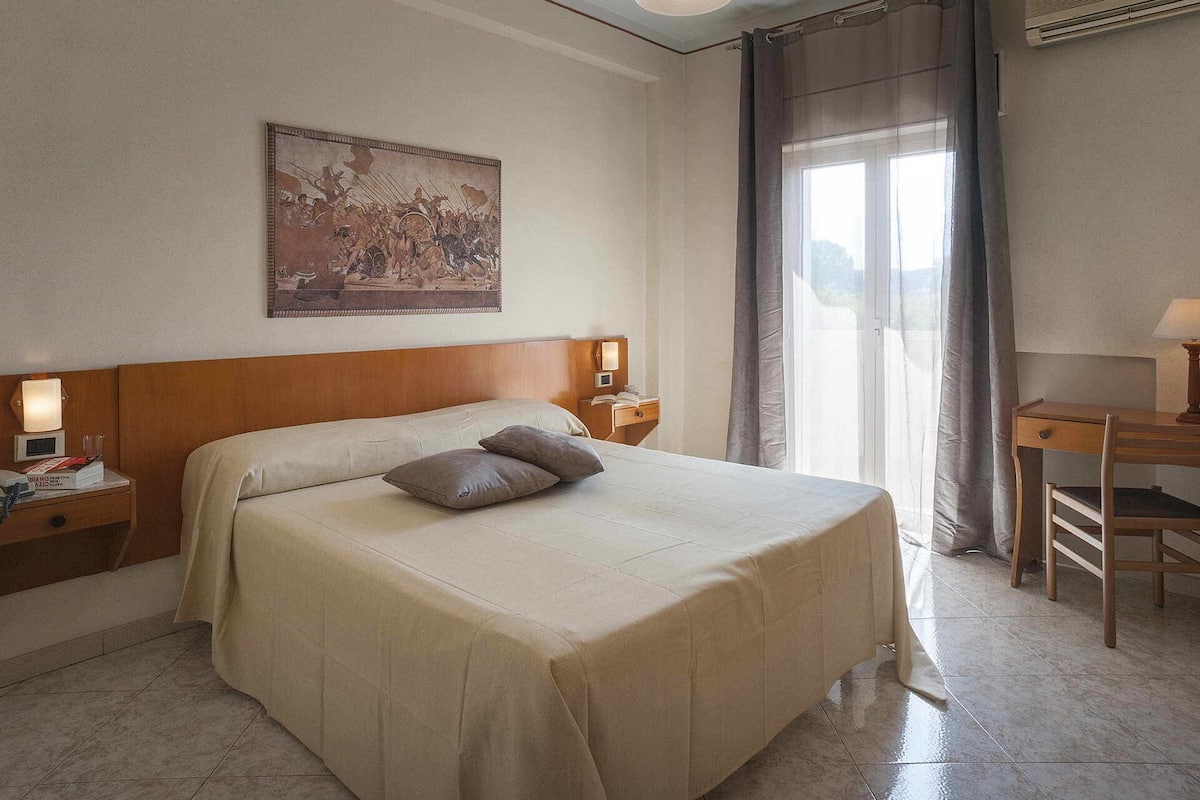 Une chambre d’hôtel avec un grand lit, un bureau, une chaise, une table de chevet avec une lampe, un tableau au mur et une fenêtre avec des rideaux.