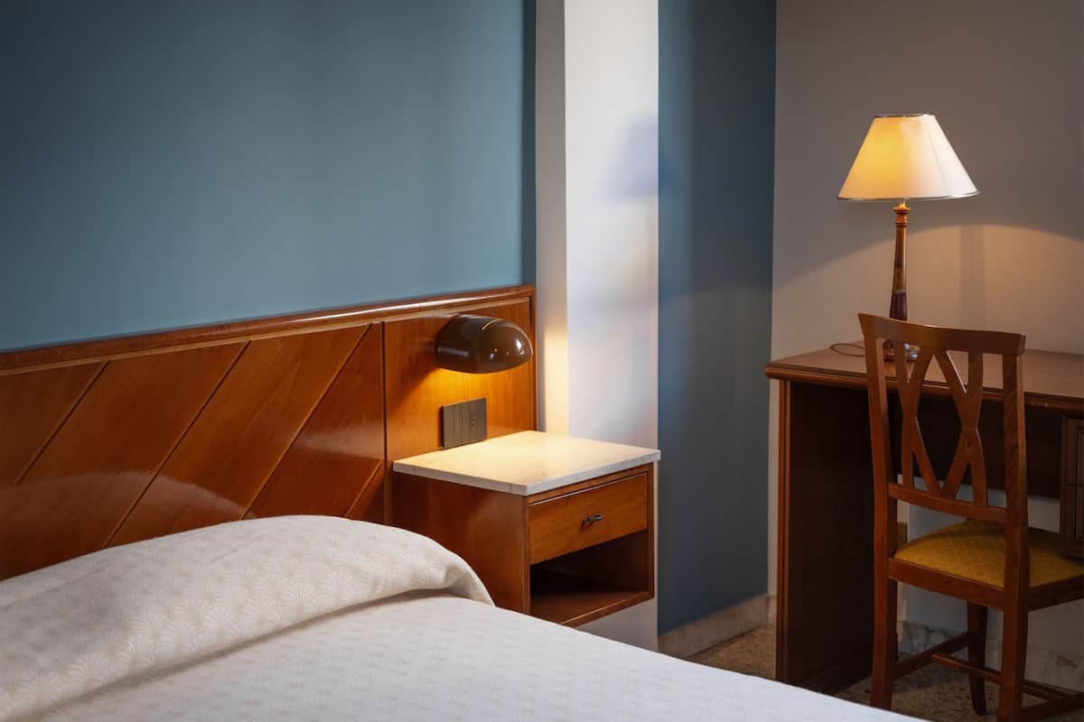 Une chambre d’hôtel avec un lit, une table de chevet avec une lampe, une chaise en bois et un bureau.
