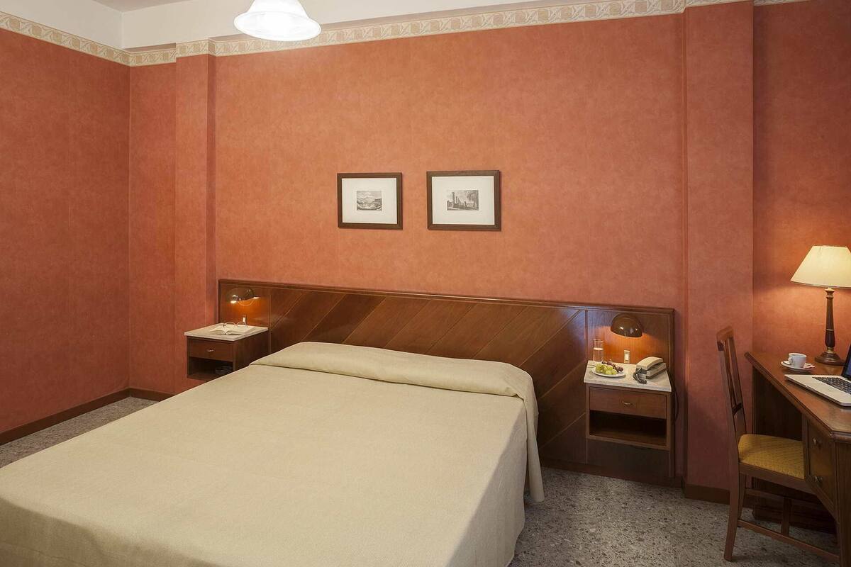 Une chambre d’hôtel avec un lit, des tables de chevet, un bureau, une chaise et deux tableaux encadrés au mur.