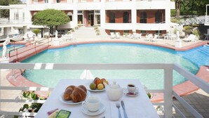 Ein Frühstückstisch am Pool mit Croissants, Früchten, Kaffee und Tee, mit Blick auf einen Pool und ein Gebäude im Hintergrund.