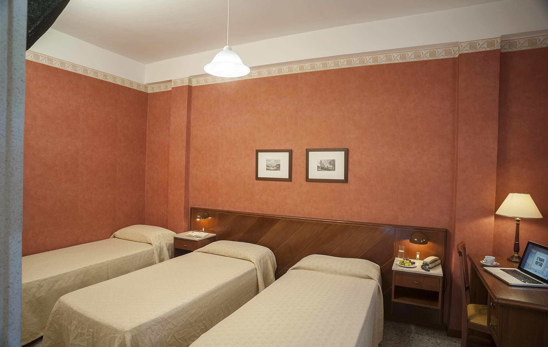 Une chambre d’hôtel avec deux lits simples, une tête de lit en bois, une table de chevet avec une lampe, un bureau avec un ordinateur portable et deux tableaux encadrés accrochés au mur.