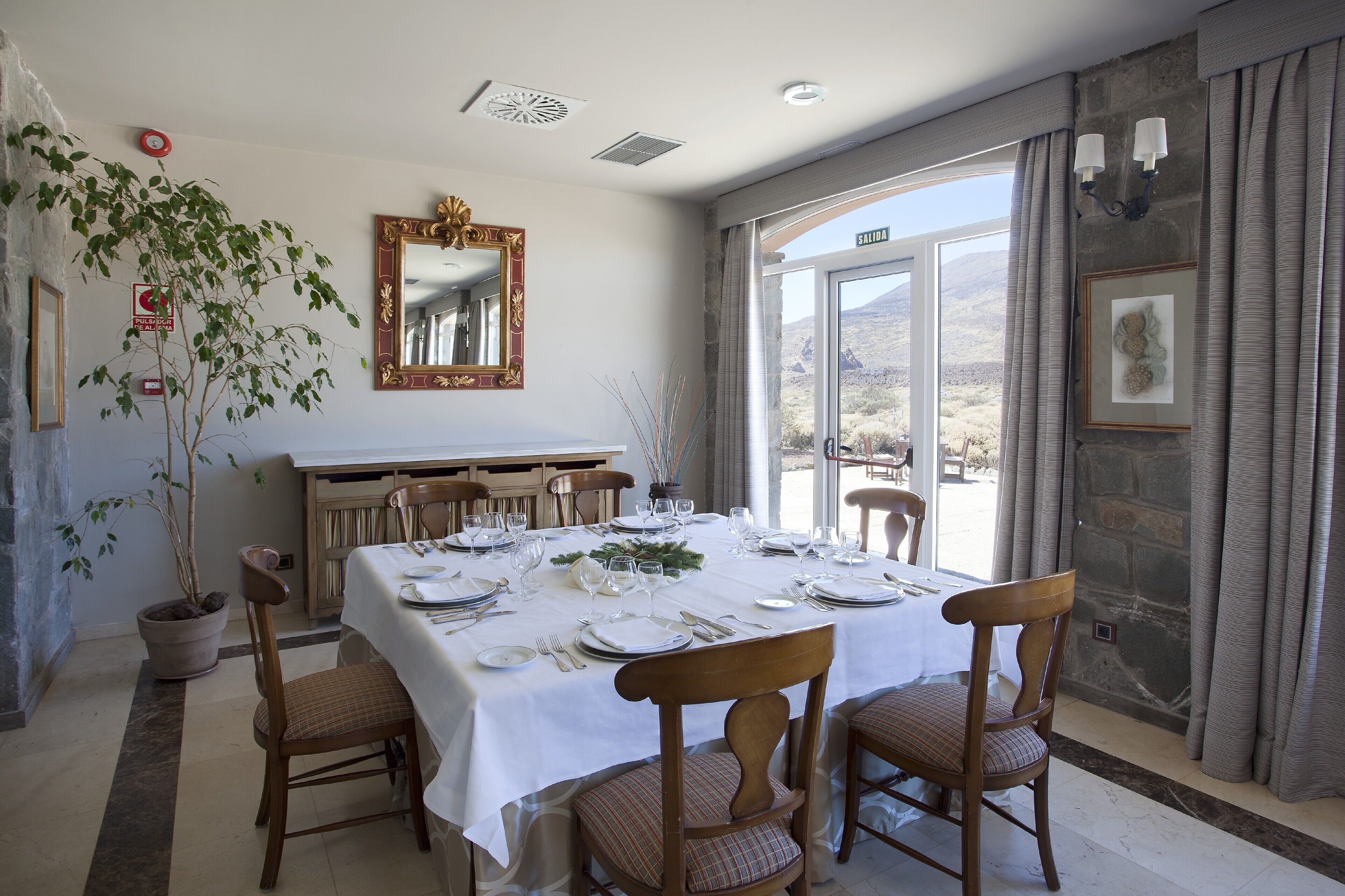 A dining area with a table set for four, a potted plant, a mirror, and a view of the outdoors.