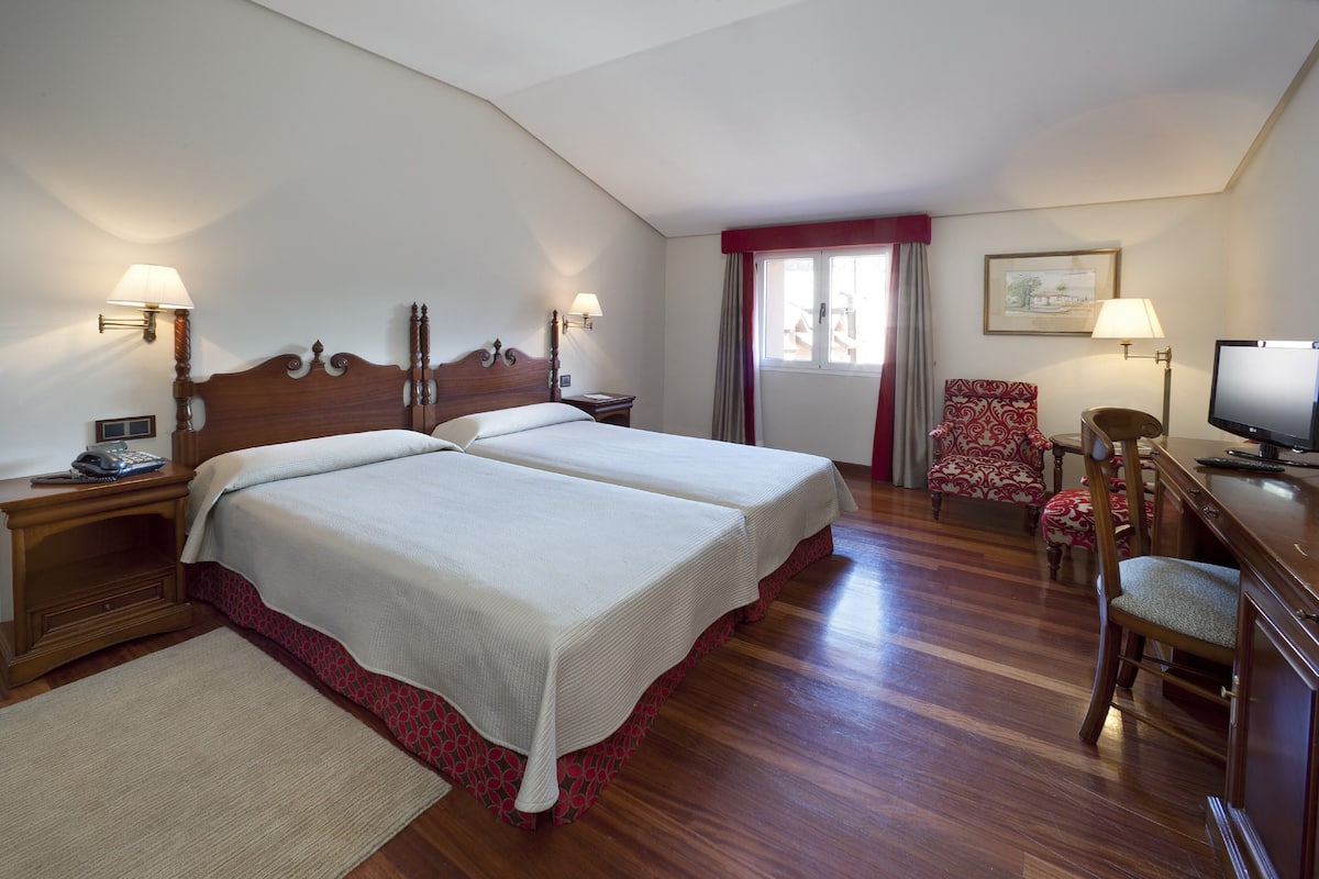 Habitación de hotel con dos camas, un escritorio con televisor, una silla y una ventana con cortinas.