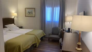 Une chambre d’hôtel avec deux lits, un bureau avec une lampe, une chaise, une fenêtre avec des rideaux et un tableau encadré au mur.