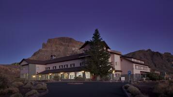 Un hotel con una gran montaña al fondo, una entrada bien iluminada y un área de estacionamiento.