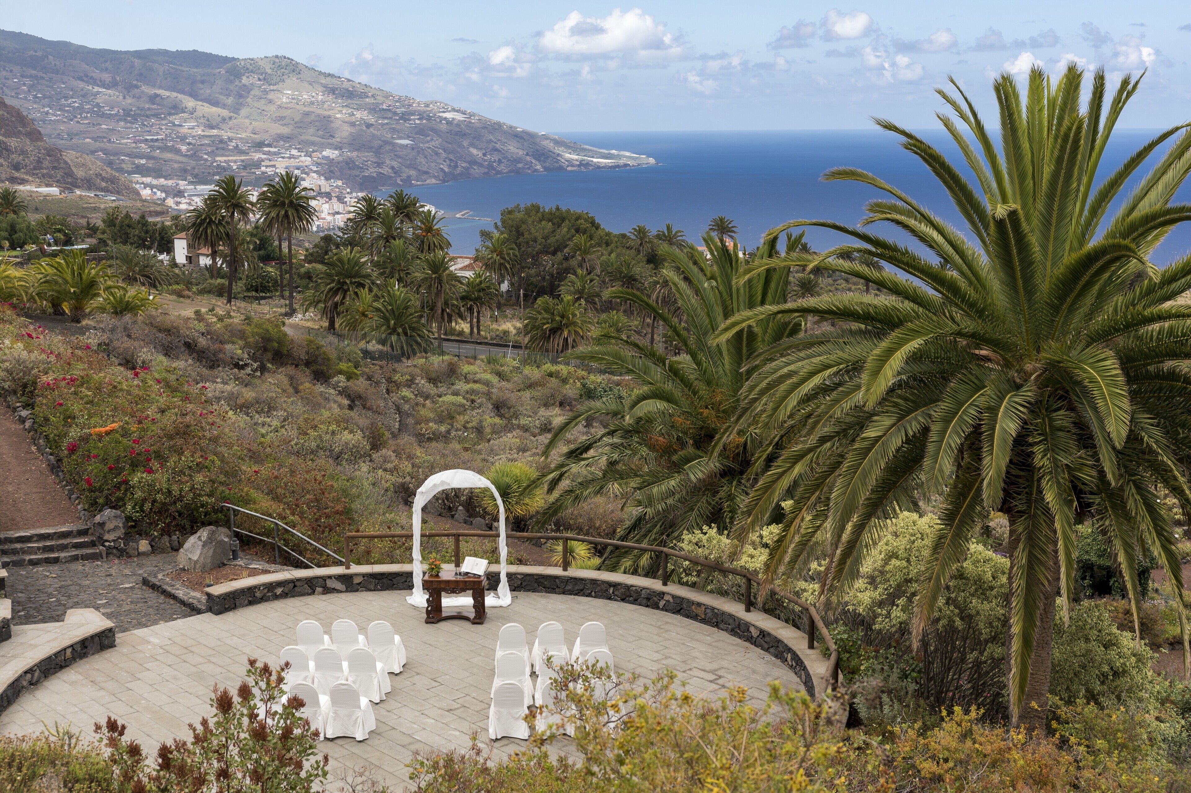 Zona relax all'aperto con sedute bianche e un tavolo in legno, circondata da palme e vista sul mare e colline in lontananza.