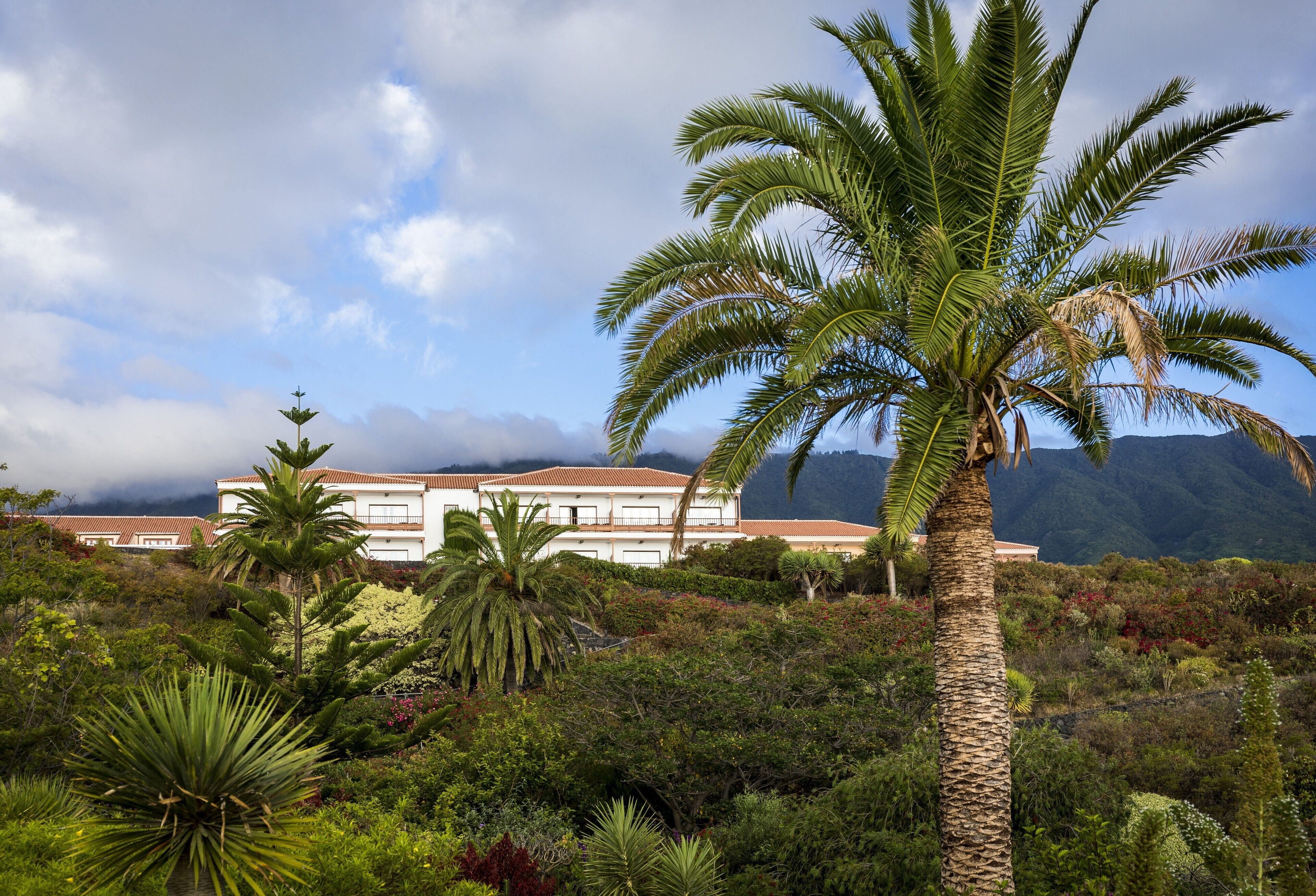 A white building with a red roof, surrounded by lush greenery and palm trees, with mountains in the background.