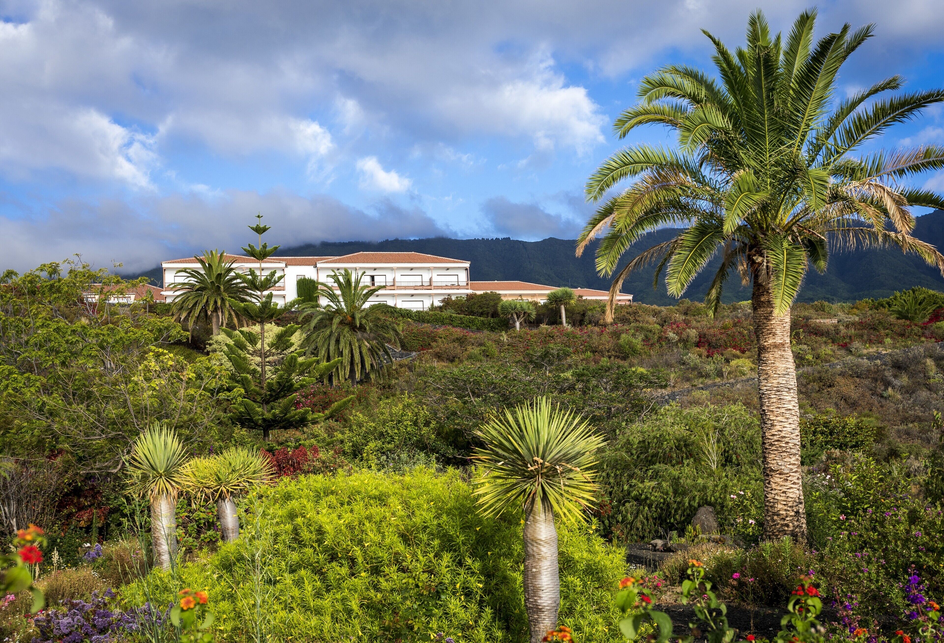A white building with a red roof surrounded by lush greenery and a mountain in the background.