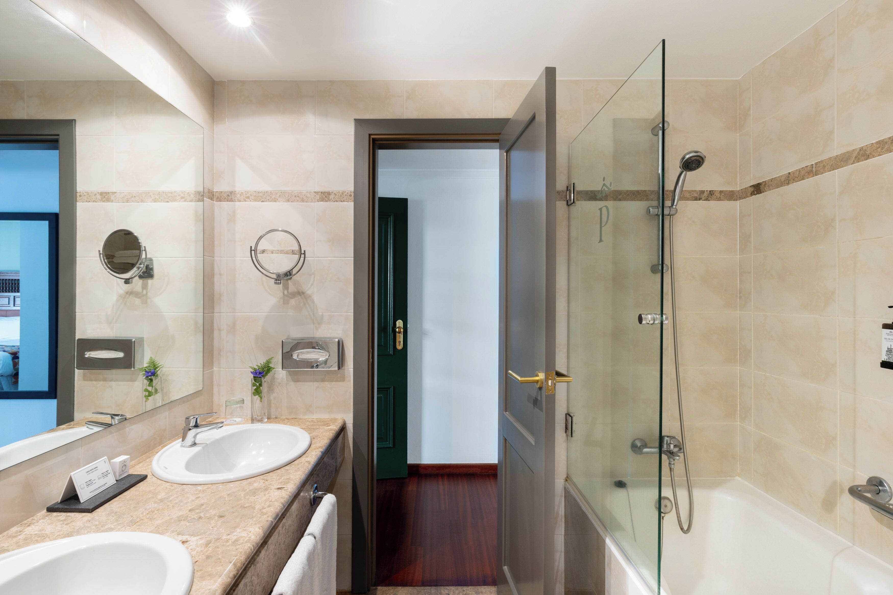 Ein modernes Badezimmer mit zwei Waschbecken, einer Duschkabine aus Glas und einer sichtbaren Tür zu einem weiteren Raum.