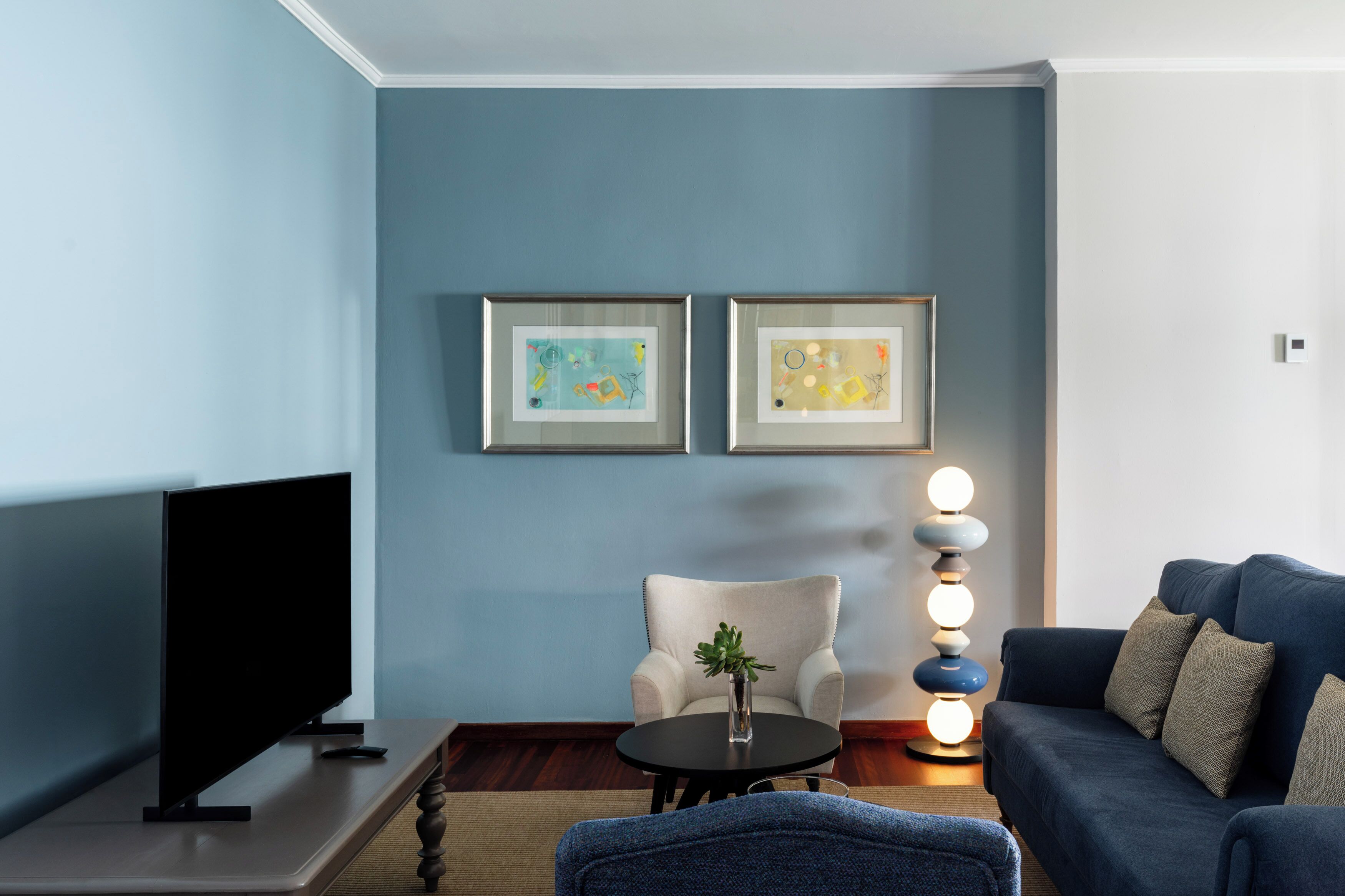 A living room with a flat-screen TV, a sofa, a chair, a small table with a plant, and two framed artworks on the wall.