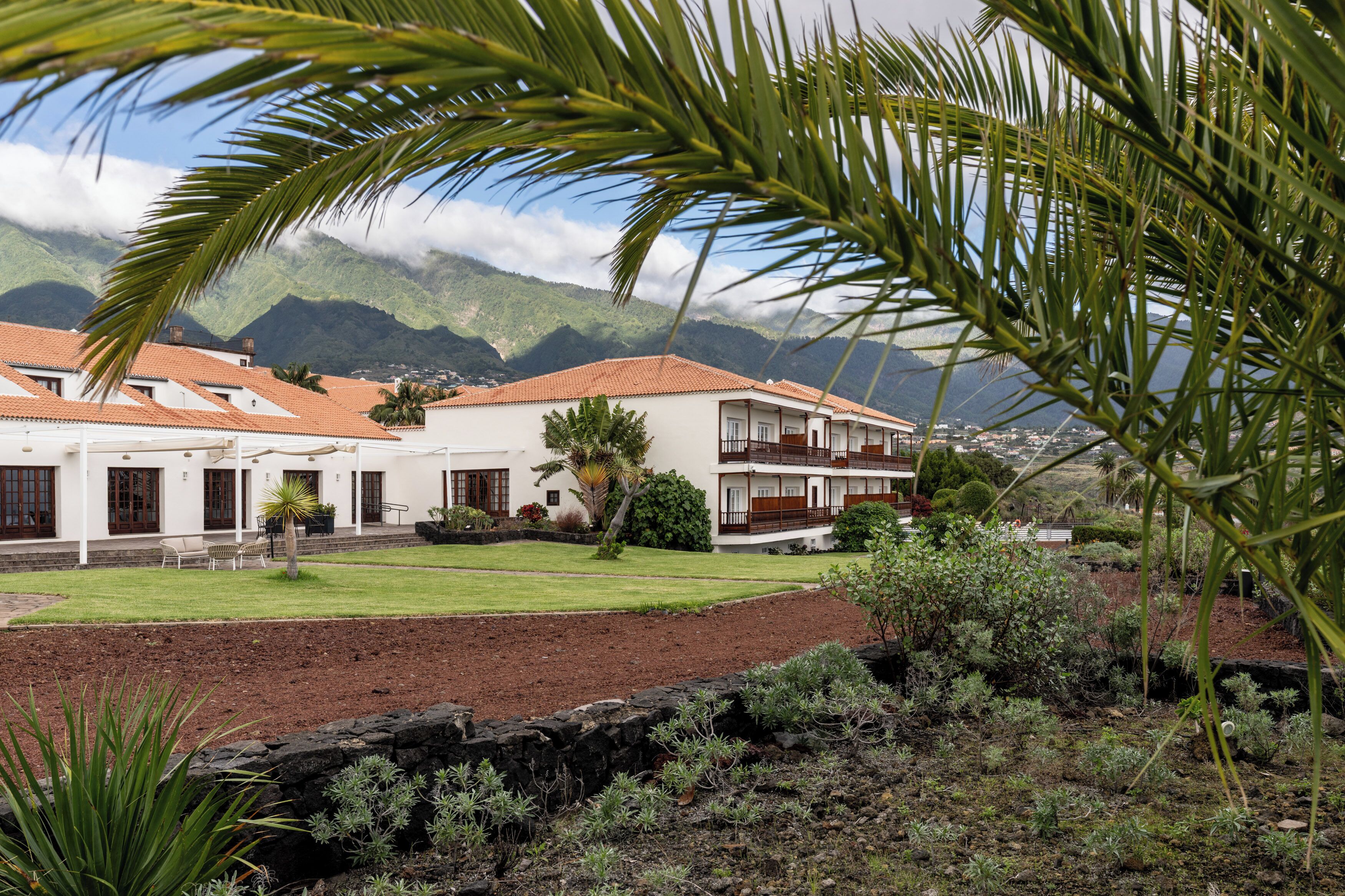 A two-story building with a red-tiled roof, surrounded by a well-maintained garden and a view of mountains in the background.