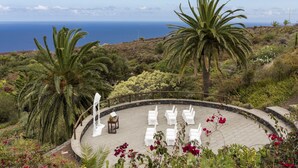 A circular outdoor seating area with white chairs and a small table, surrounded by lush greenery and a view of the ocean. - Parador de La Palma (Brena Baja)
