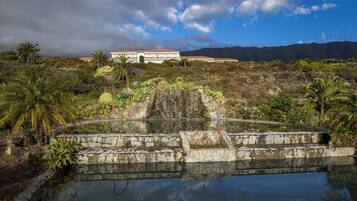 Un puente de piedra sobre un estanque que refleja el paisaje, con vegetación tropical, un edificio grande al fondo y montañas a lo lejos.
