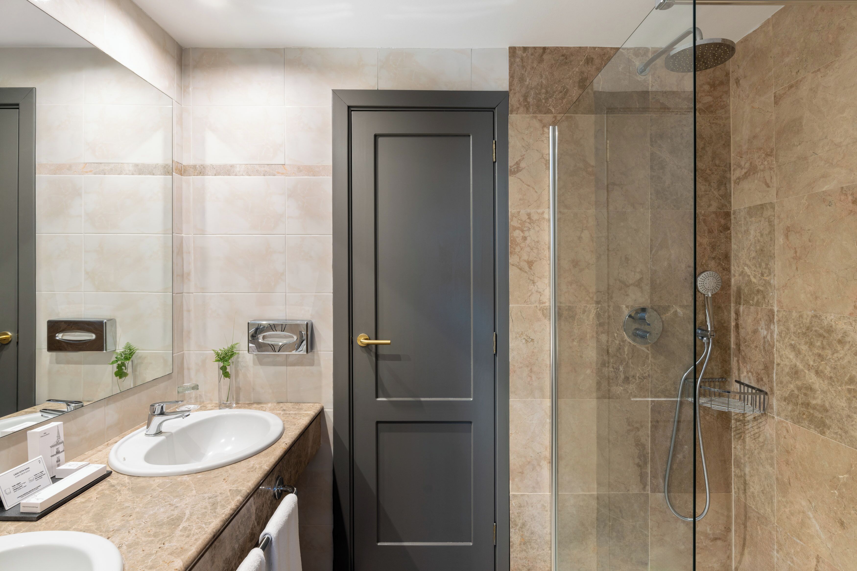 Un baño moderno con ducha envuelta en vidrio, una puerta oscura y encimera de mármol con lavamanos y espejo.