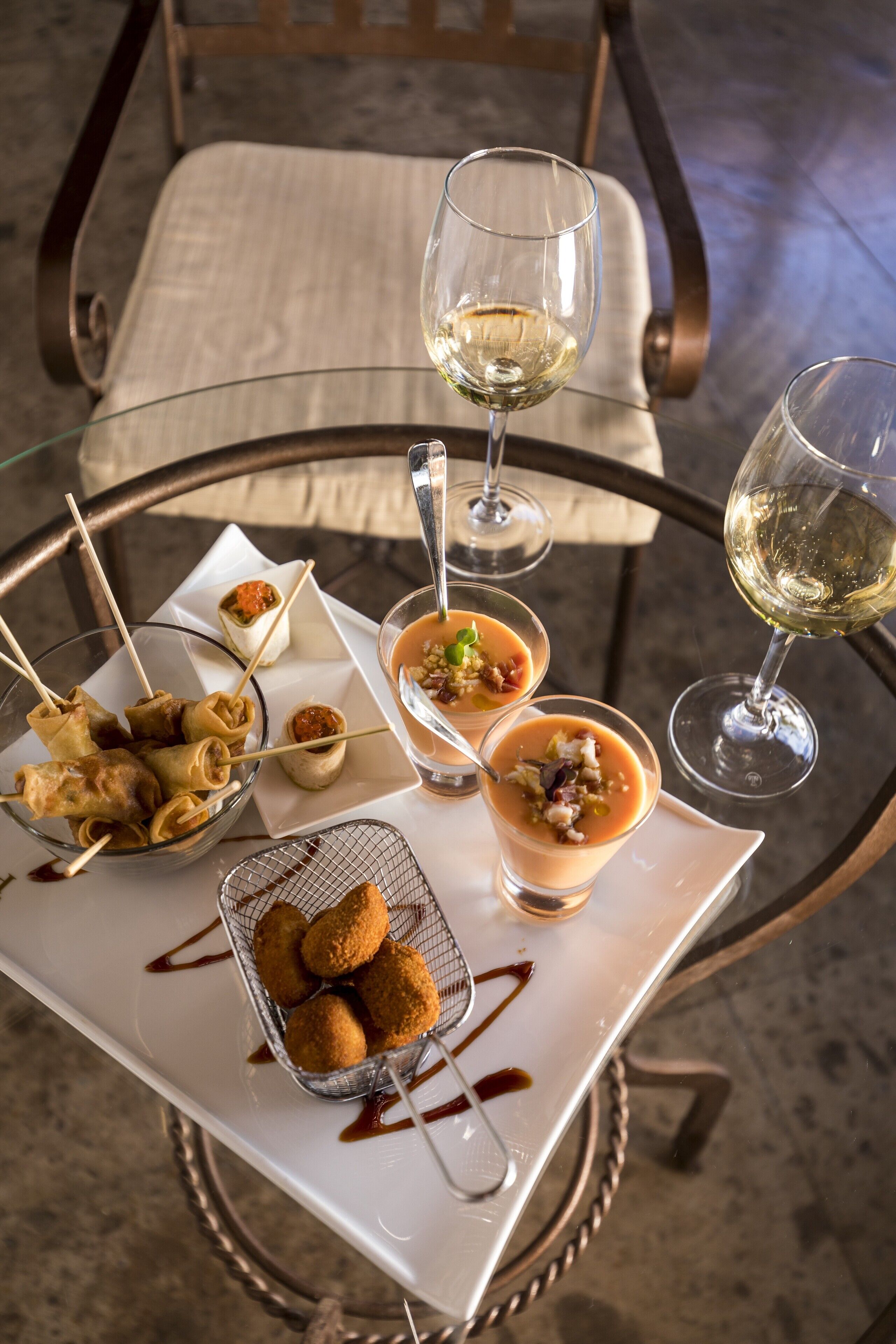 Una mesa con diferentes platos, copas de vino blanco y una silla.