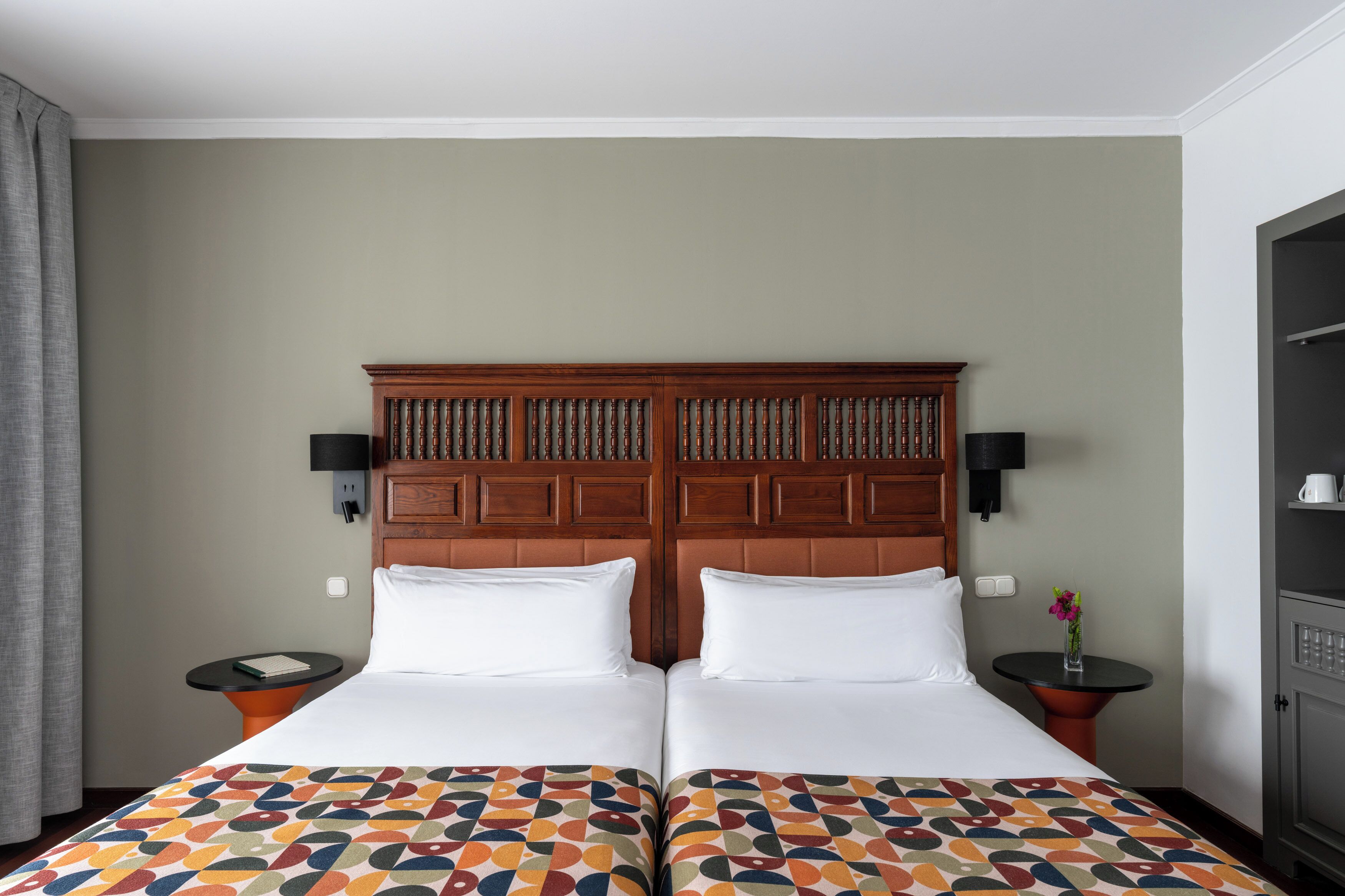 Ein Doppelbett mit gemustertem Bettbezug, ein Holz-Kopfteil, zwei Nachttische jeweils mit einer Lampe sowie ein an der Wand befestigter Fernseher.