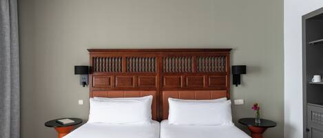 Ein Doppelbett mit gemustertem Bettbezug, ein Holz-Kopfteil, zwei Nachttische jeweils mit einer Lampe sowie ein an der Wand befestigter Fernseher.