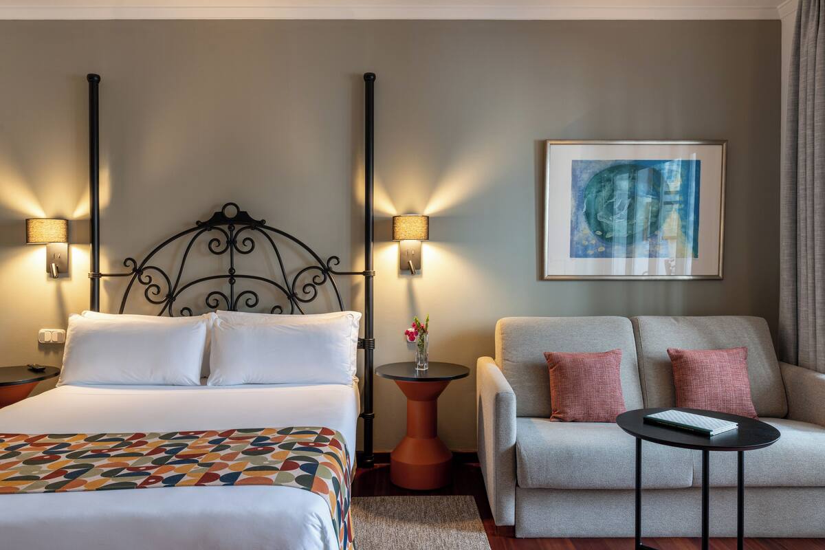 Habitación de hotel con cama, dos lámparas de noche, un sofá, una mesita y un cuadro enmarcado en la pared.