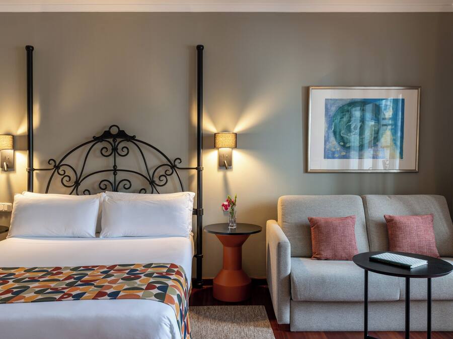 A hotel room with a bed, two bedside lamps, a sofa, a small table, and a framed artwork on the wall.