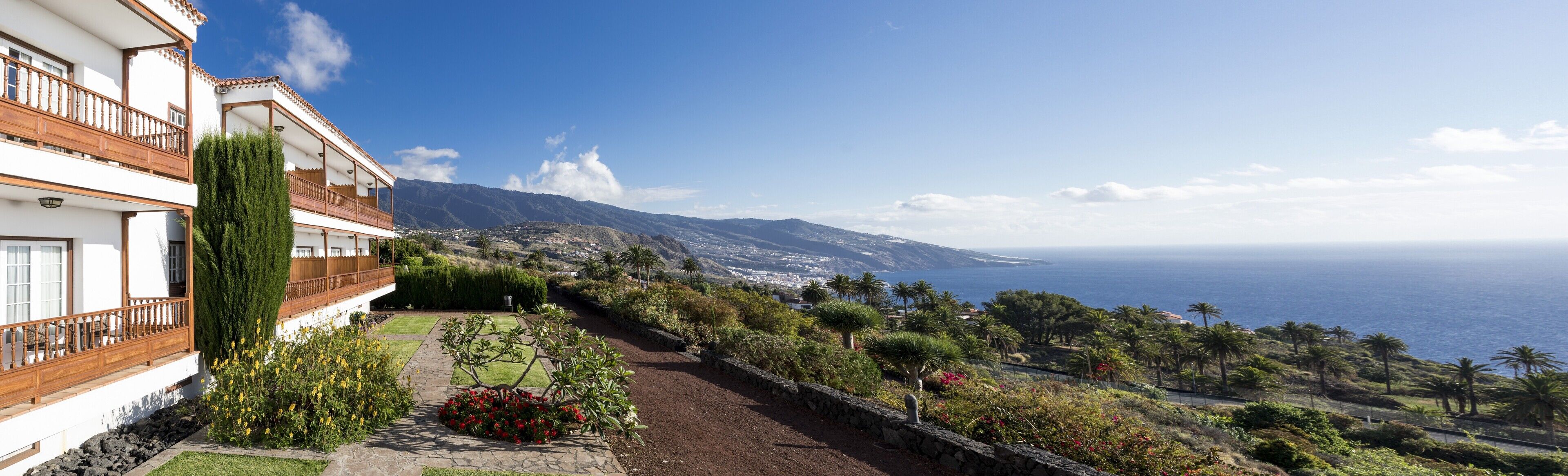 A coastal property with a balcony overlooking a sea view, a garden with various plants, and a mountainous backdrop.