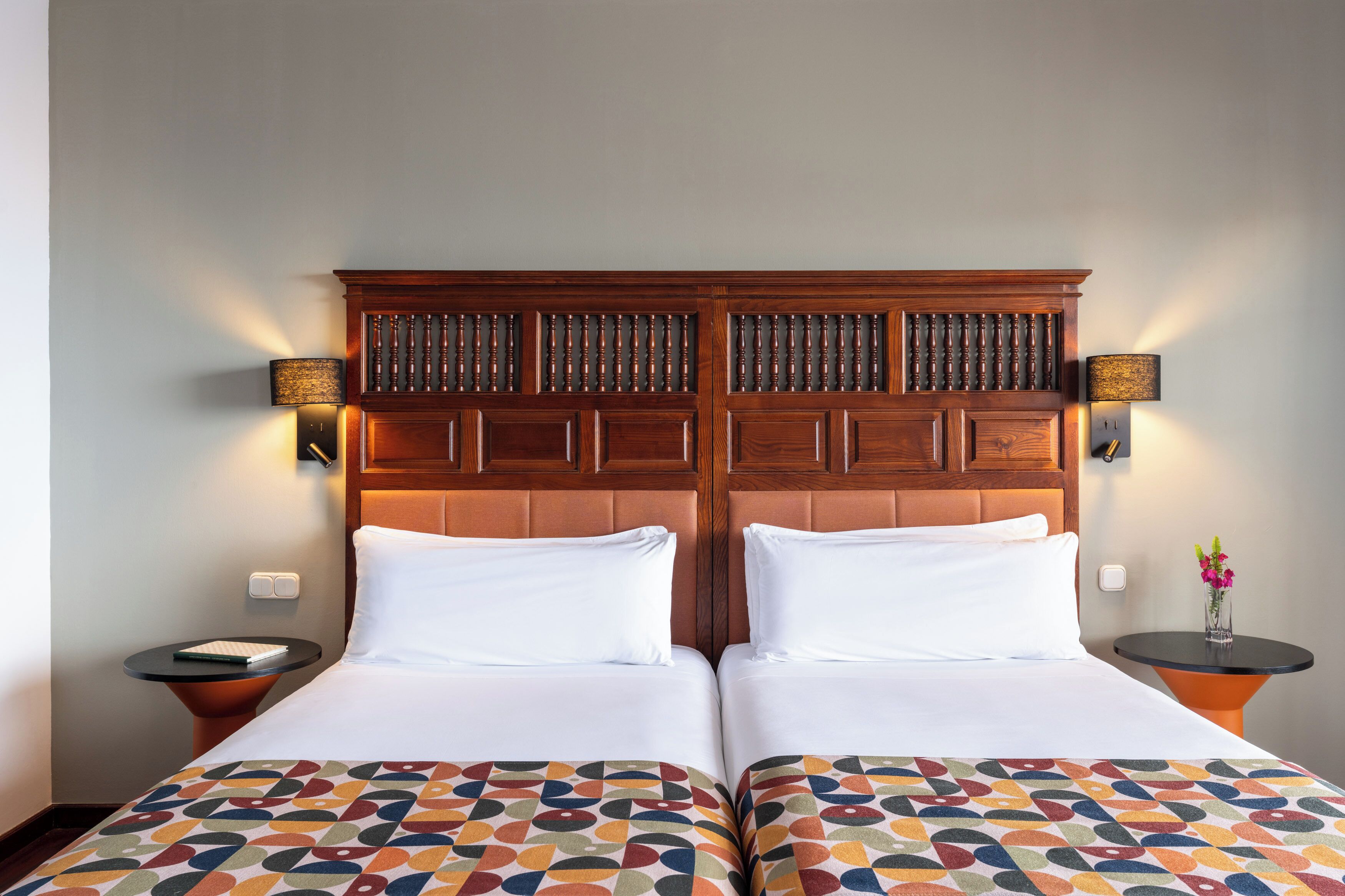 A double bed with a patterned bedspread, a wooden headboard, two bedside tables with lamps, and a small table with a vase of flowers.