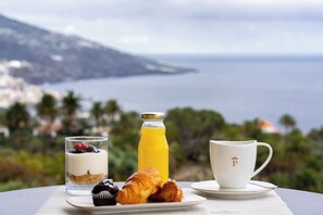 A breakfast setting with a glass of yogurt parfait, a croissant, a muffin, a glass of orange juice, and a white cup with a logo, all on a table with a scenic view of the sea and mountains in the background.
