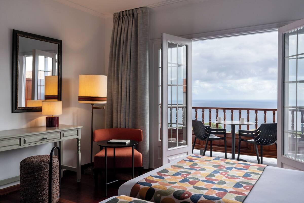Habitación de hotel con cama, escritorio, silla, mesa con sillas, balcón con vistas y ventana.