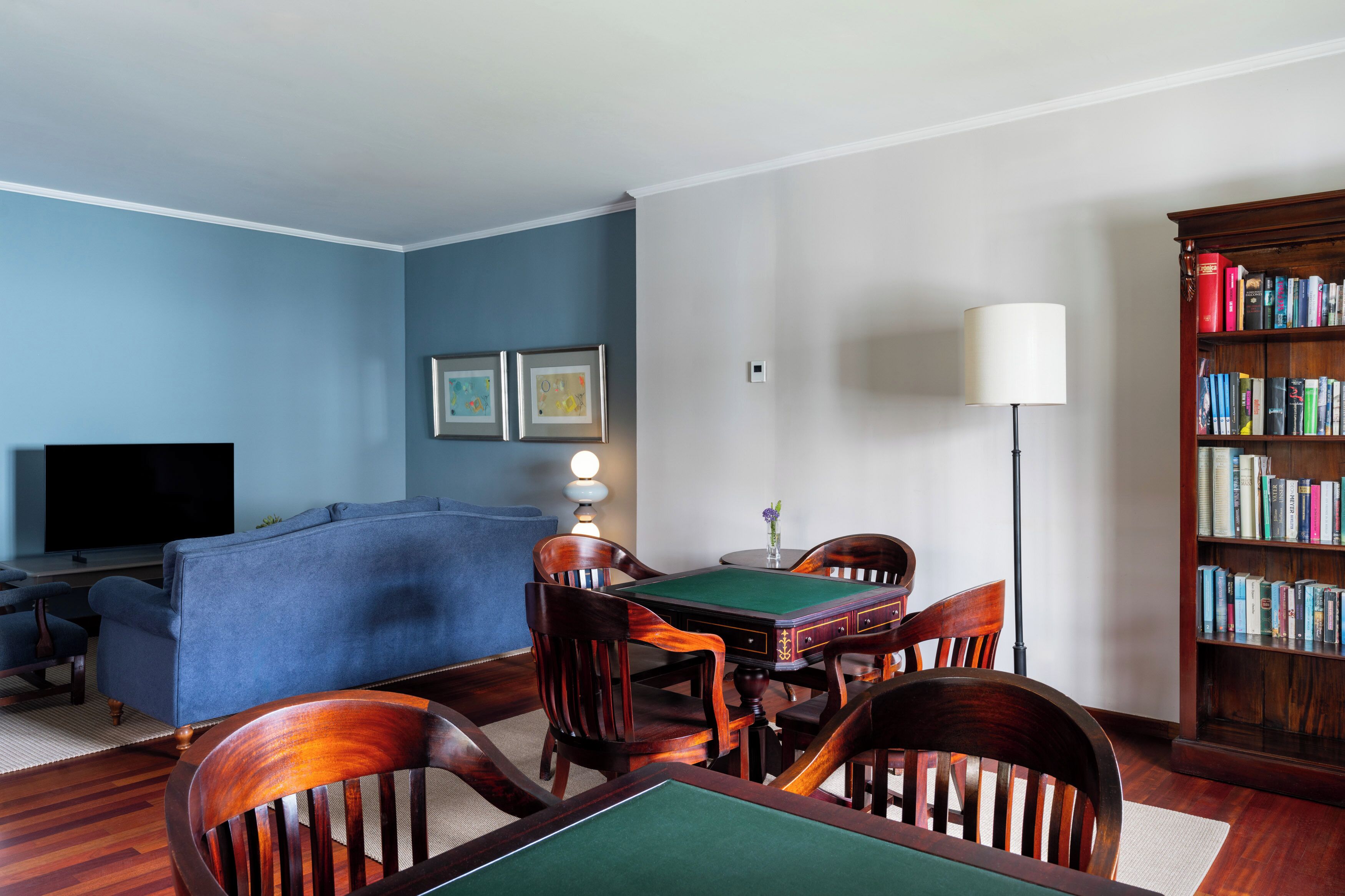 A living room with a blue sofa, wooden chairs, a green table, a bookshelf, and a lamp.