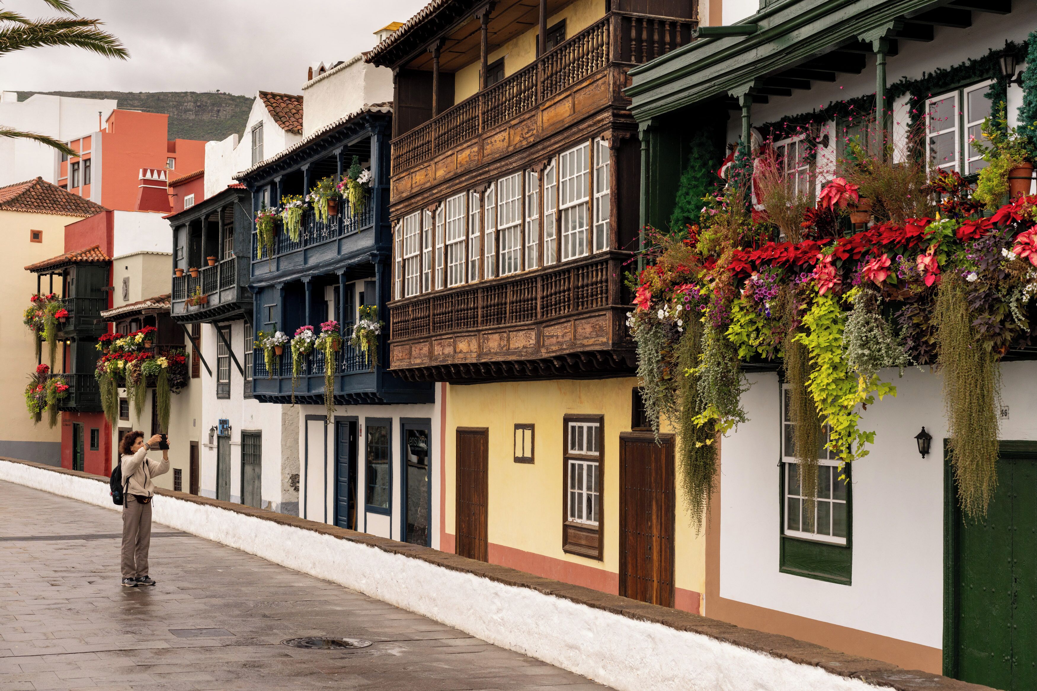 A street with traditional buildings, balconies with hanging plants, and a person taking a photo.