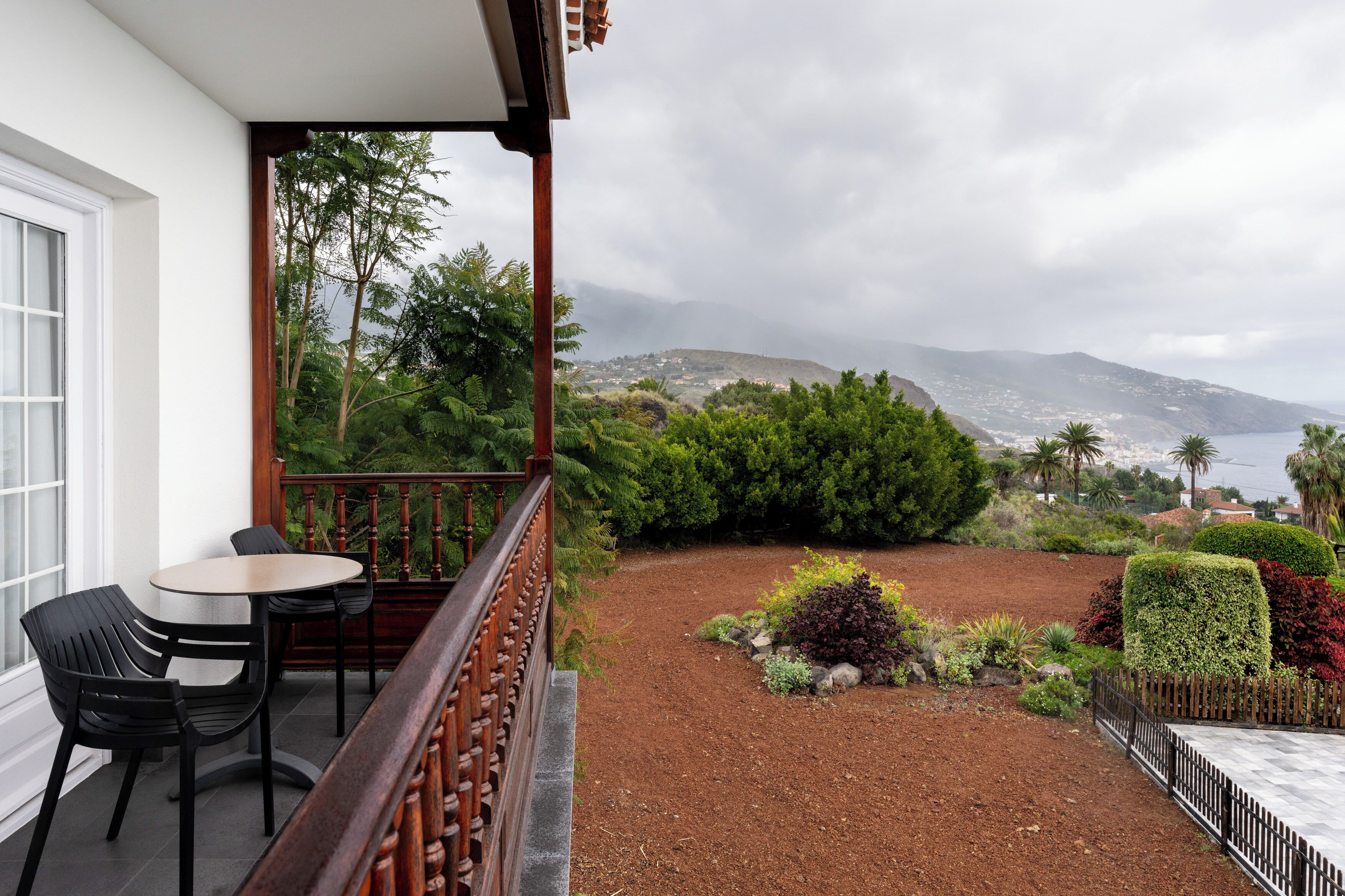 Un balcon avec une table et des chaises, une vue sur un jardin aménagé et des montagnes au loin.