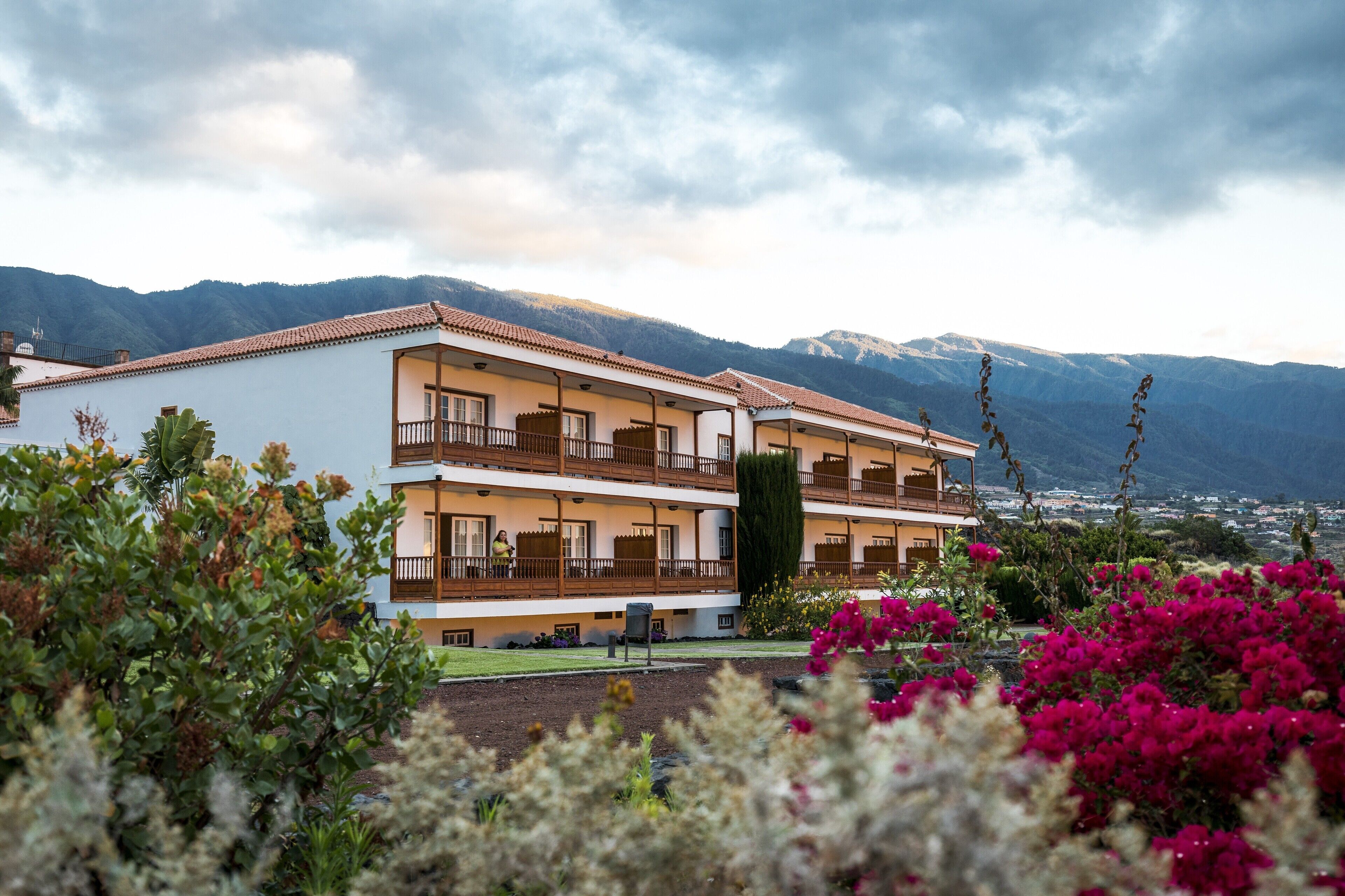Un hotel de dos pisos con balcones, rodeado de montañas y plantas en flor.