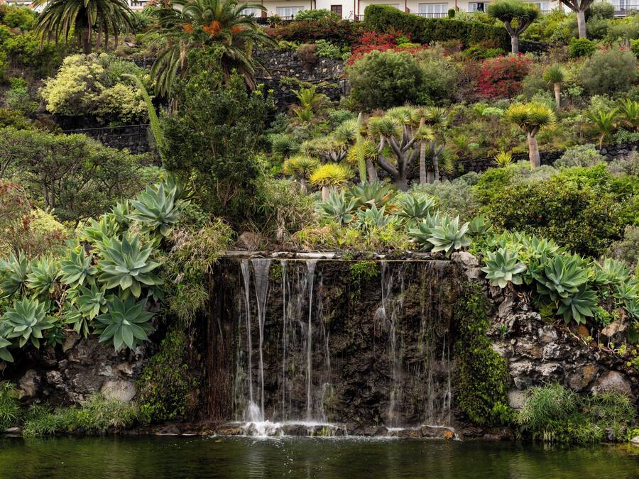 Una cascada que desemboca en un estanque rodeado de exuberante vegetación y un edificio grande con tejado rojo al fondo.